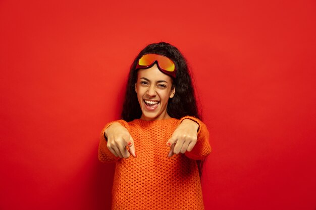 African-american young brunette woman's portrait in ski mask on red studio background. Concept of human emotions, facial expression, sales, ad, winter sport and holidays. Pointing on, laughting.