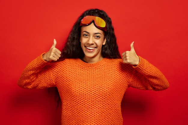 African-american young brunette woman's portrait in ski mask on red studio background. Concept of human emotions, facial expression, sales, ad, winter sport and holidays. Smiles and thumb up.