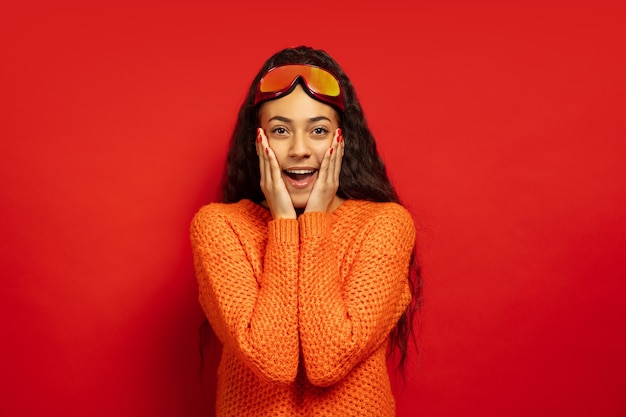 African-american young brunette woman's portrait in ski mask on red studio background. Concept of human emotions, facial expression, sales, ad, winter sport and holidays. Surprised, astonished.