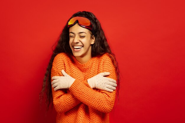 Портрет афро-американской молодой брюнетки в лыжной маске на красном фоне студии. Концепция человеческих эмоций, выражения лица, продаж, рекламы, зимних видов спорта и праздников. Согревание от холода, смех.
