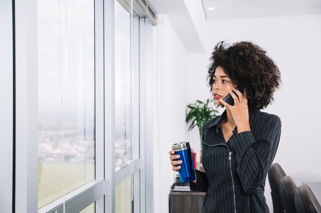 african american young lady with thermos talking on smartphone near window