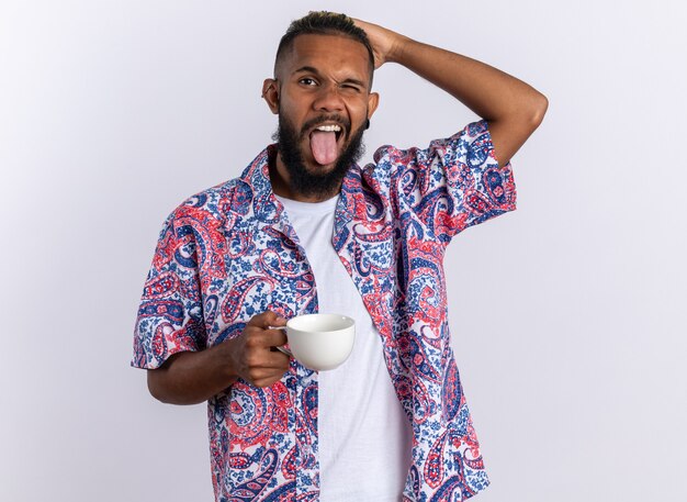 African american young man in colorful shirt holding cup happy and joyful sticking out tongue standing over white background