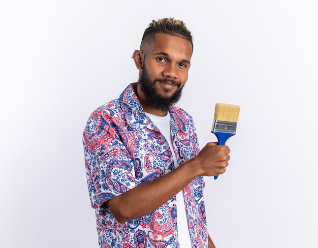 African american young man in colorful shirt holding paint brush looking at camera smiling