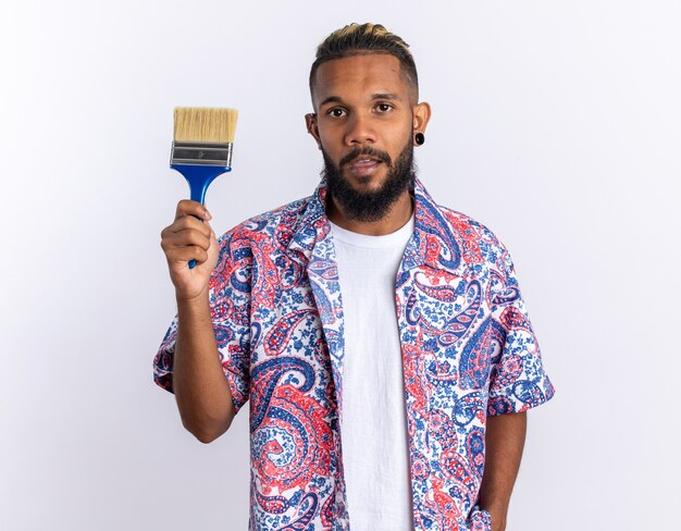 African american young man in colorful shirt holding paint brush looking at camera with serious face standing over white