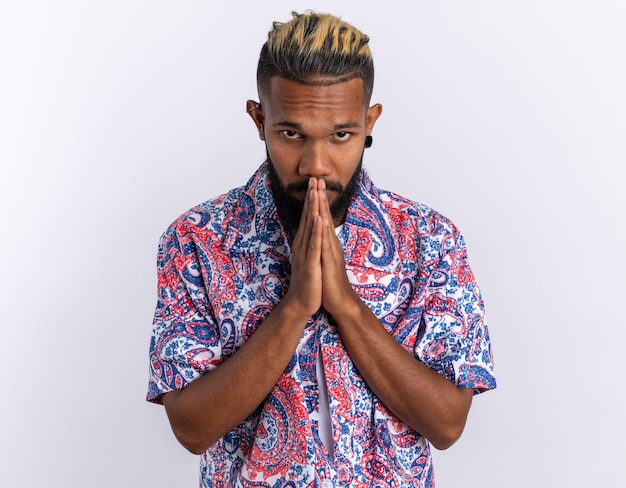 African american young man in colorful shirt holding palms together like praying standing over white