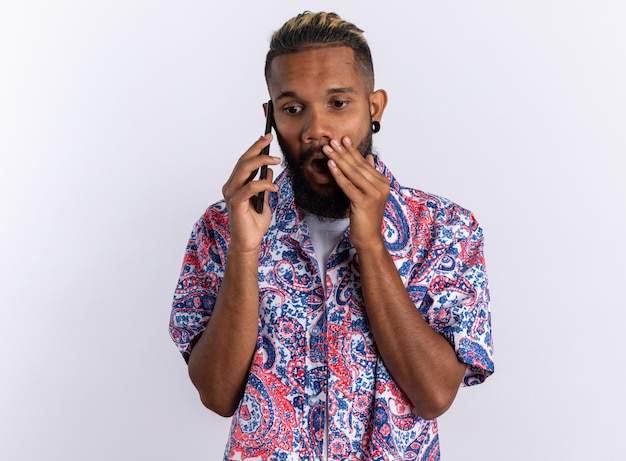 African american young man in colorful shirt looking amazed and shocked while talking on mobile phone standing over white