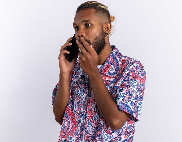 African american young man in colorful shirt looking amazed while talking on mobile phone standing over white