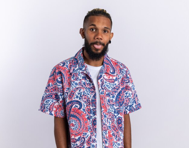 African american young man in colorful shirt looking at camera happy and positive sticking out tongue