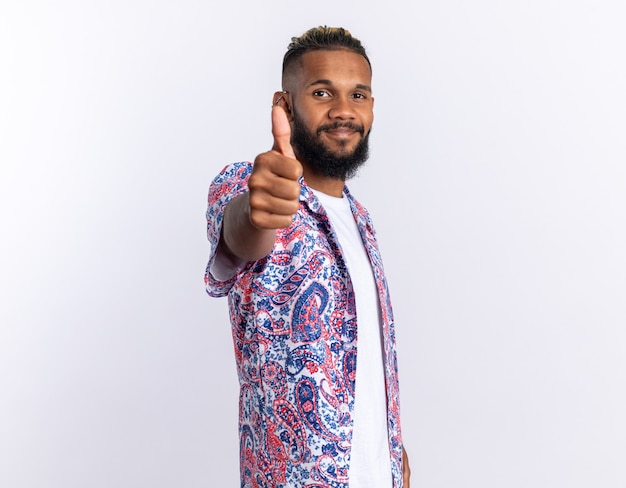 African american young man in colorful shirt looking at camera smiling cheerfully showing thumbs up standing over white