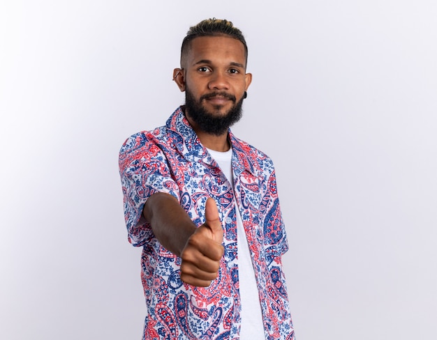 African american young man in colorful shirt looking at camera smiling confident showing thumbs up standing over white background