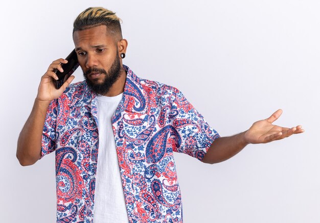 Афро-американский молодой человек в красочной рубашке выглядит смущенным во время разговора по мобильному телефону, стоя на белом фоне