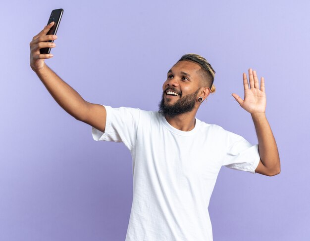 手で手を振って幸せで陽気な笑顔のスマートフォンを使用して自分撮りをしている白いTシャツのアフリカ系アメリカ人の若い男