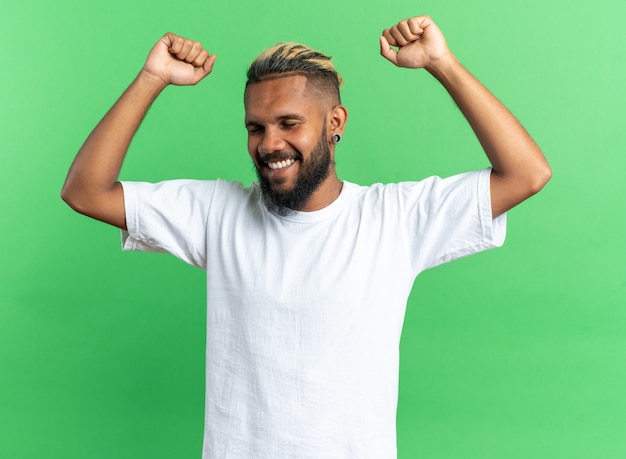 緑の背景の上に立って彼の成功を喜んで幸せで興奮した握りこぶしを握りしめる白いTシャツのアフリカ系アメリカ人の若い男