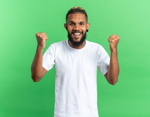 Афро-американский молодой человек в белой футболке счастлив и взволнован, сжимая кулаки, радуясь своему успеху, стоя на зеленом фоне