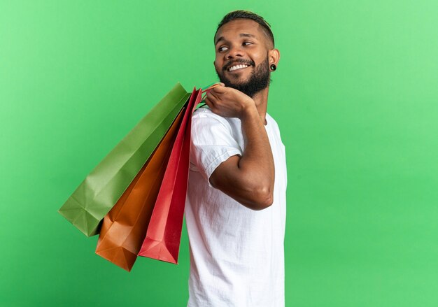 African american young man in white t-shirt holding paper bags looking aside smiling cheerfully happy and positive standing over green background