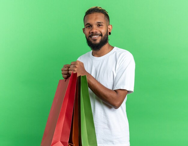 Афро-американский молодой человек в белой футболке держит бумажные пакеты, весело улыбаясь, глядя в камеру