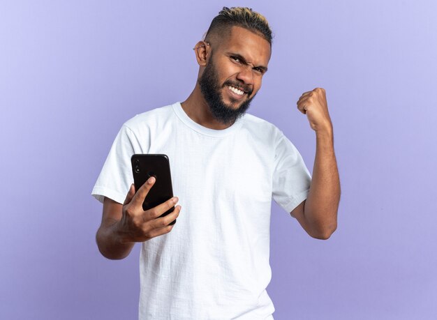 Афро-американский молодой человек в белой футболке держит смартфон, сжимая кулак, счастлив и взволнован, радуясь своему успеху, стоя на синем фоне