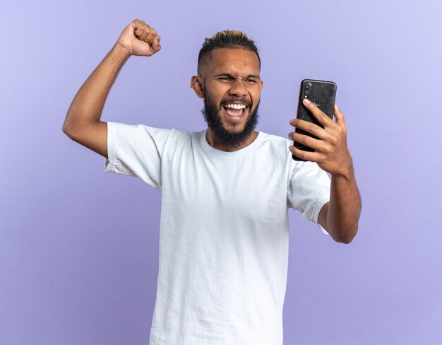 スマートフォンを握りこぶしを握って幸せな白いTシャツのアフリカ系アメリカ人の若い男