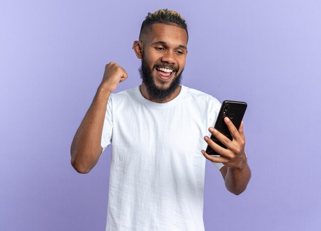 スマートフォンを握りこぶしを握って幸せな白いTシャツのアフリカ系アメリカ人の若い男