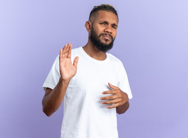 African american young man in white t-shirt looking at camera confused and displeased making stop gesture with hands standing over blue background