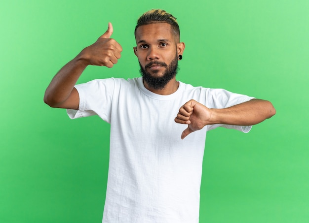 African american young man in white t-shirt looking at camera confused showing thumbs up