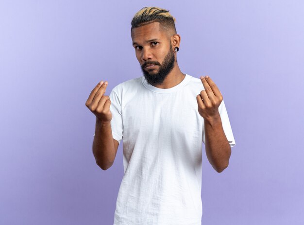 Афро-американский молодой человек в белой футболке смотрит в камеру, делая деньги жестом, потирая пальцы, стоя на синем фоне