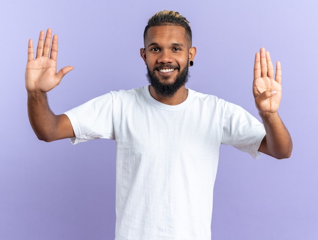 African american young man in white t-shirt looking at camera smiling cheerfully showing number nine standing over blue background