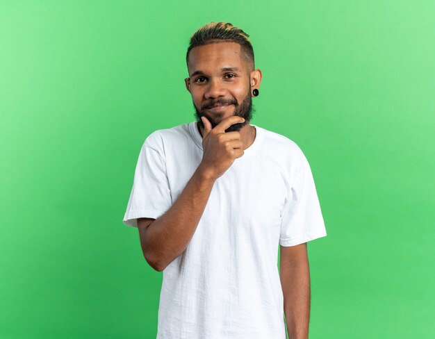 흰색 티셔츠를 입은 아프리카계 미국인 청년이 턱에 손을 대고 녹색 배경 위에 서서 즐겁게 웃고 있는 카메라를 바라보고 있다
