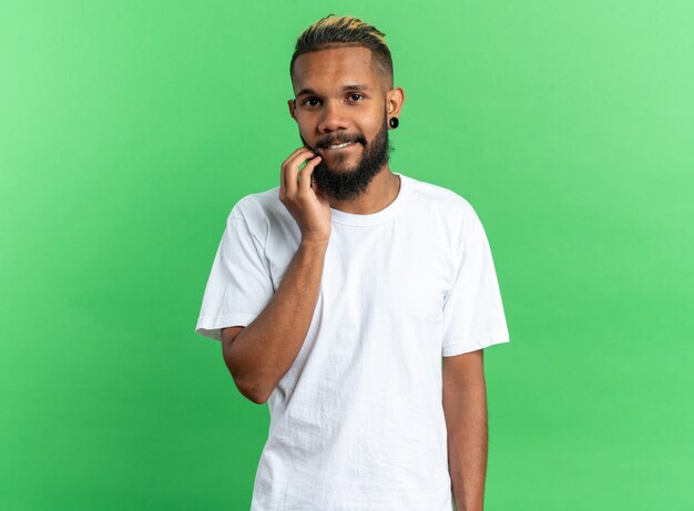Афро-американский молодой человек в белой футболке смотрит в камеру с рукой на подбородке, дружелюбно улыбаясь, стоя на зеленом фоне