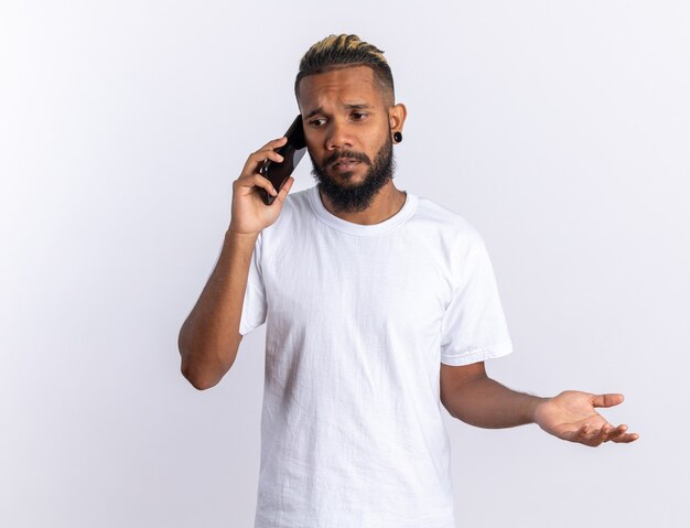 African american young man in white t-shirt looking confused while talking on mobile phone standing over white background