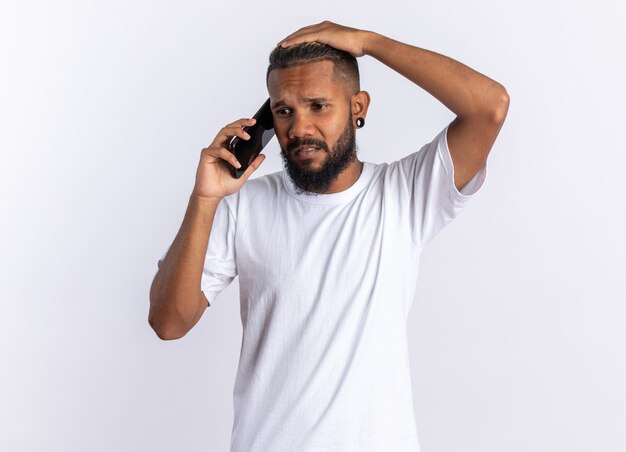 흰색 위에 서있는 휴대 전화에 얘기하는 동안 혼란 찾고 흰색 티셔츠에 아프리카 계 미국인 젊은 남자