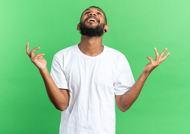 African american young man in white t-shirt looking up happy and excited raising arms standing over green background