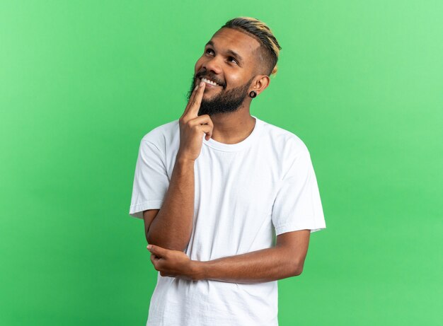 Афро-американский молодой человек в белой футболке смотрит вверх озадаченно улыбаясь, стоя над зеленым