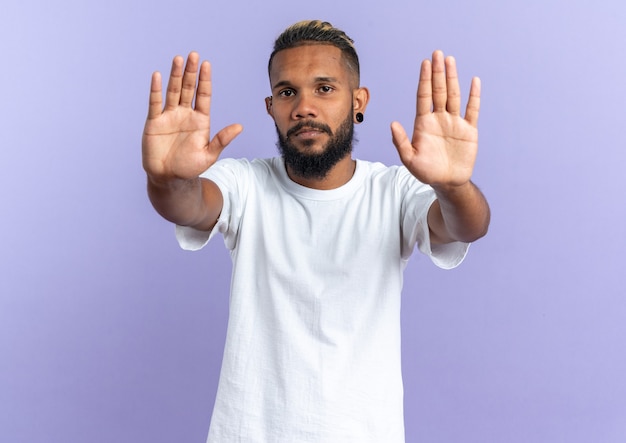 Афро-американский молодой человек в белой футболке смотрит в камеру с серьезным лицом, делая стоп-жест