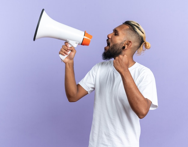 African american young man in white t-shirt shouting to megaphone happy and excited standing over blue