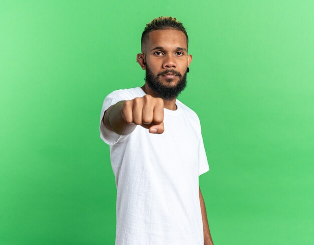 African american young man in white t-shirt showing fist at camera looking with serious confident expression standing over green background