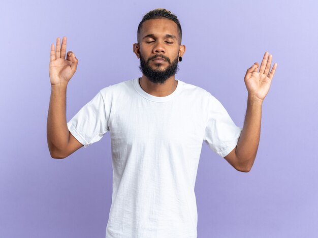 African american young man in white t-shirt trying to relax making meditation gesture with fingers with eyes closed