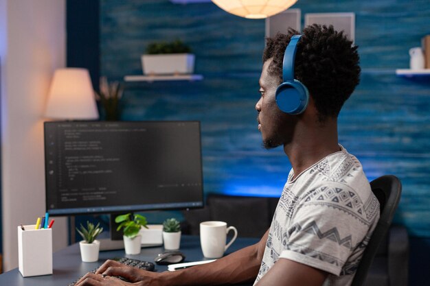 African american young programmer wearing headphone while typing security codes for business website working rmeote from home. Employee programming software application. Software on screen