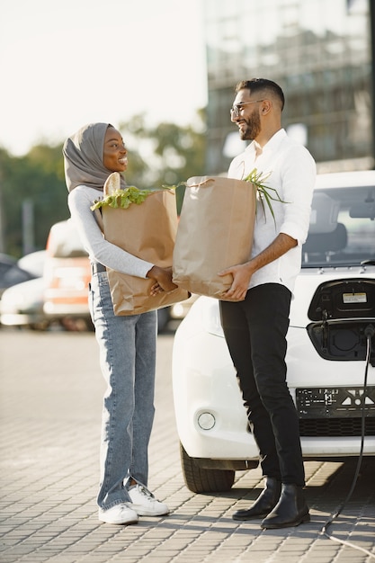 Африканская арабская пара стоит с продуктами возле электромобиля. Зарядка электромобиля на АЗС