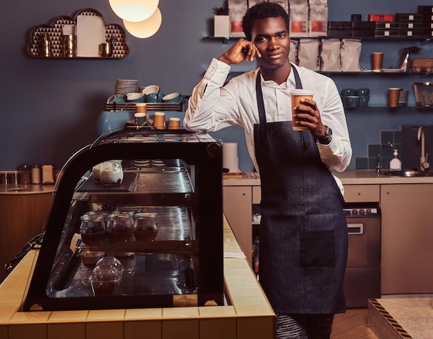 African barista smiling at camera relaxing after workday with coffee while leaning on counter at coffee shop.