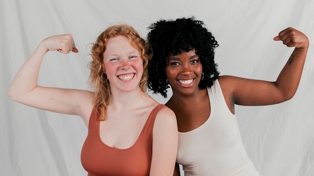 An african and blonde young woman flexing his muscle against grey backdrop