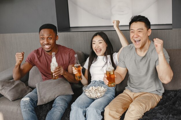 African boy and Asian couple clink a bottles with a beer. Friends watching football game, eating popcorn. People rooting for a football team.