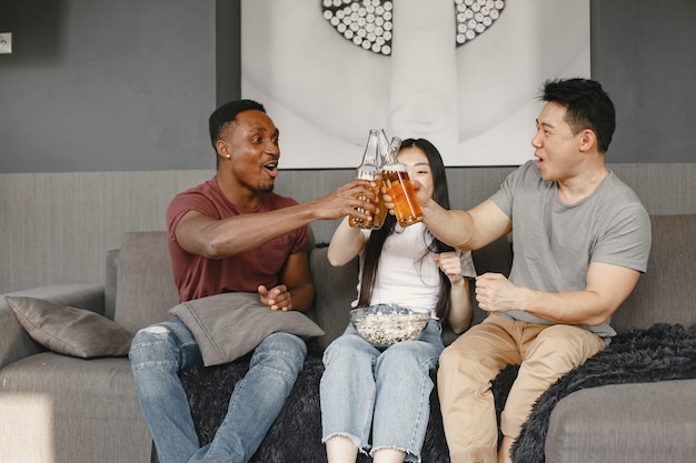 アフリカの少年とアジアのカップルがビールを飲みながらボトルをカチッと鳴らす サッカーの試合を見ながらポップコーンを食べる友達