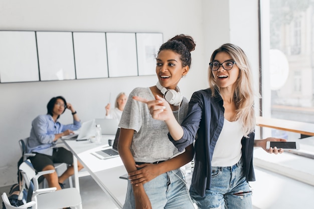 Free photo african business-lady in casual attire looking away while talking with blonde colleague in glasses. portrait of asian programmer working with laptop and black curly girl.