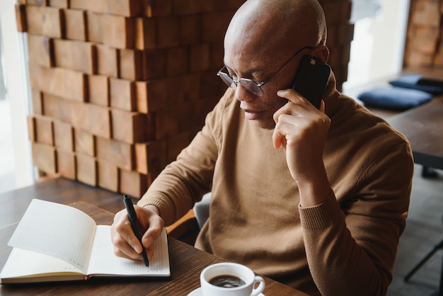 カフェ​の​テーブル​に​座って​電話​で​話している​アフリカ​の​ビジネスマン​、​モバイル​で​話している​ラップ​トップ​の​紙​と​コーヒー​ハウス​で​遠く​で​働いている​忙しい​起業家​、​カフェテリア​で​昼食​を​とっている​黒人​男性
