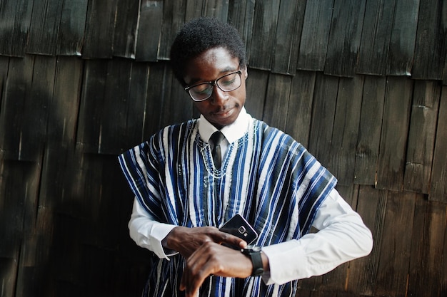 African businessman in traditional clothes and glasses with mobile phone at hand looking at his watches against wooden wall