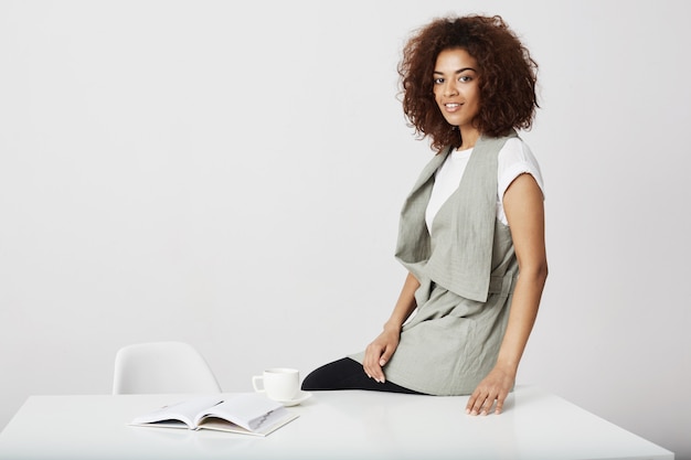 African businesswoman smiling sitting on table at workplace over white wall.