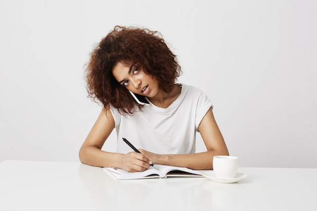 African businesswoman talking on phone at workplace over white wall. Copy space.