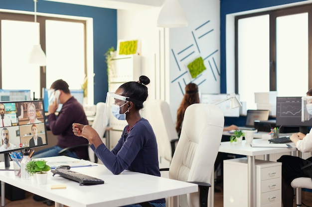 African businesswoman wearing wireless headphones during online call with business people wearing face mask. New normal business office Multiethnic team working respecting social distance during globa