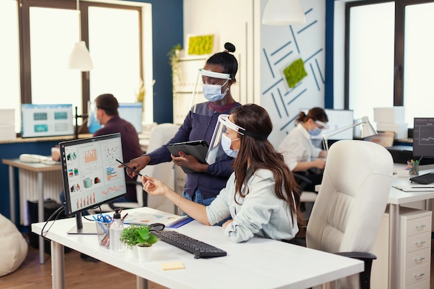 African and caucasian coworkers working together wearing face mask against covid19. Diverse team working respecting social distancing during global pandemic with coronavirus. New normal.